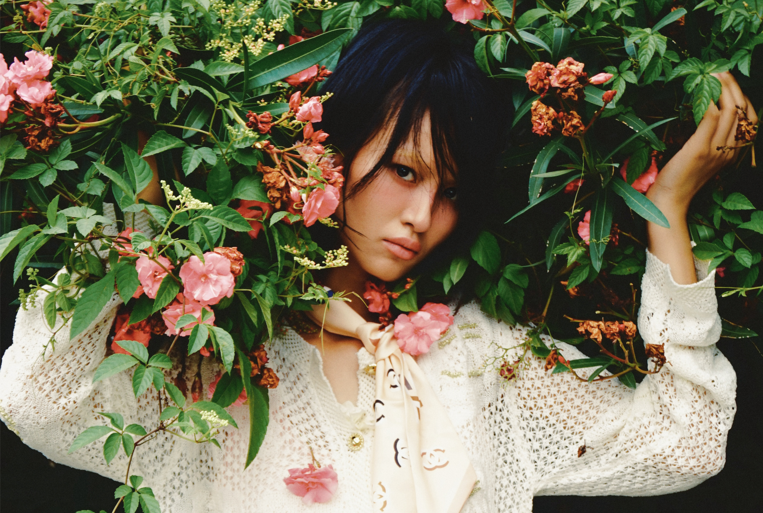 Person in floral setting wearing white knit sweater and scarf, surrounded by leaves.