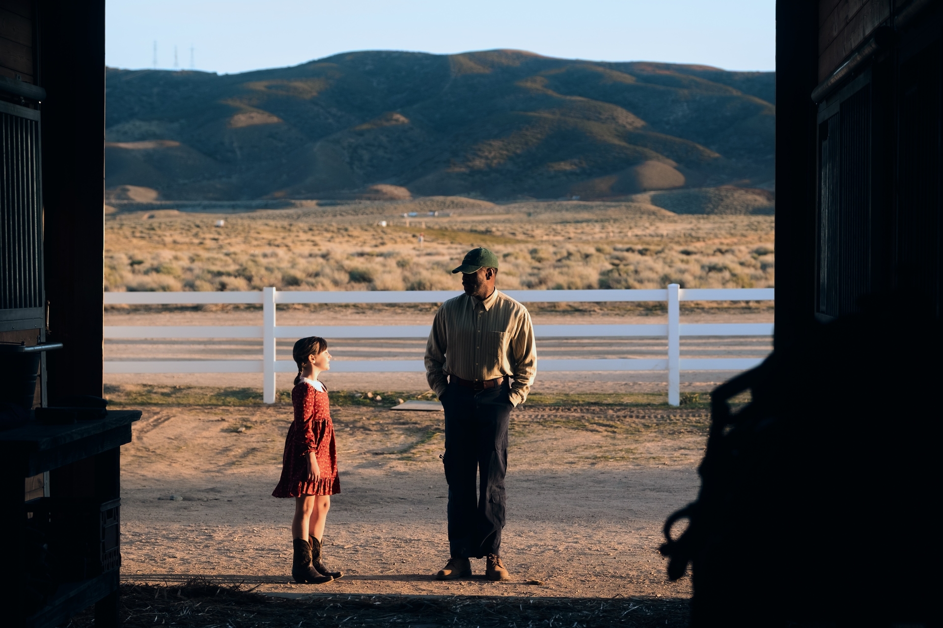 Child and adult conversing in a barn doorway against a scenic mountain backdrop.