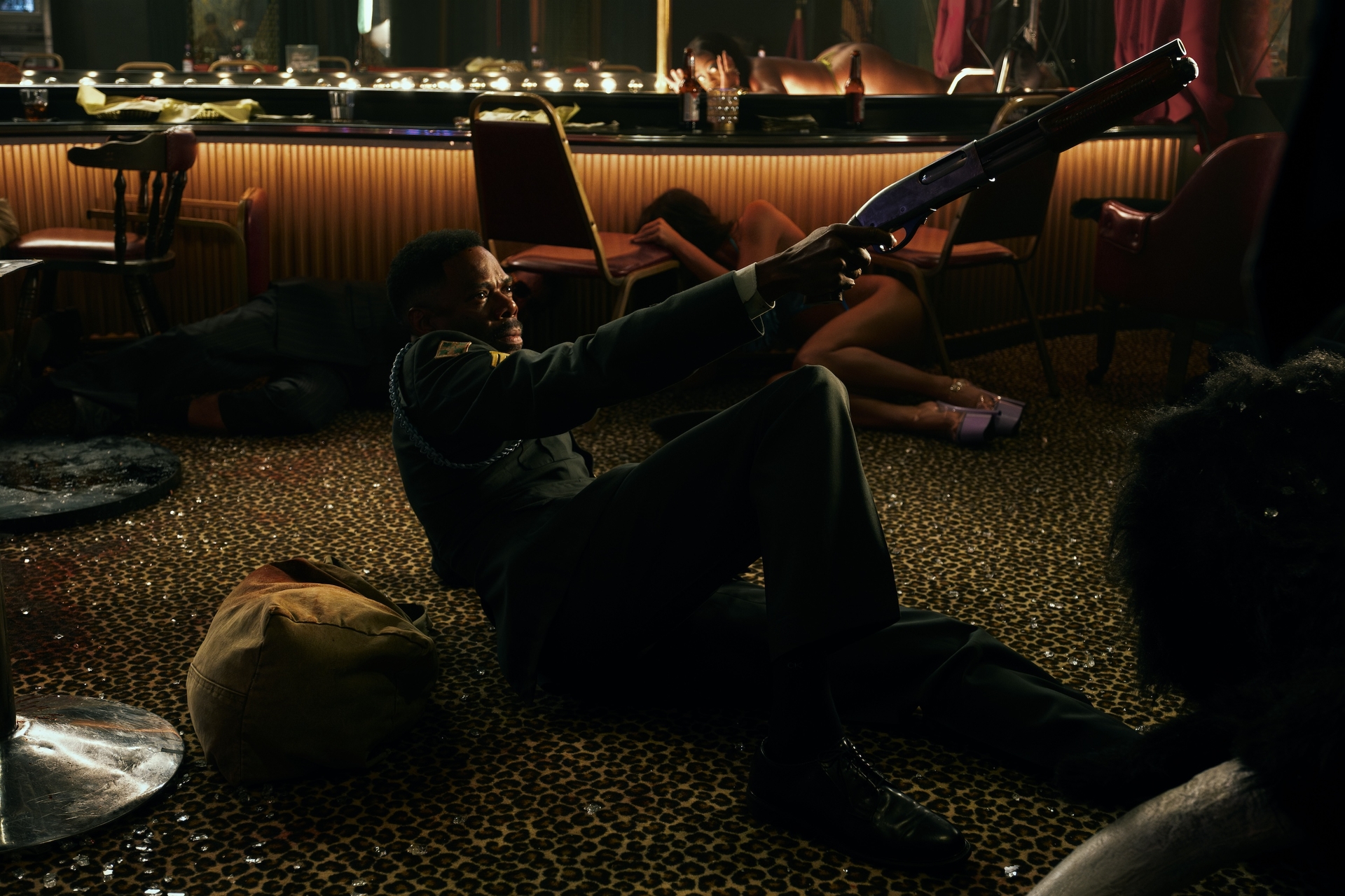 Man holding shotgun in dramatic nightclub scene with fallen chairs and scattered people.