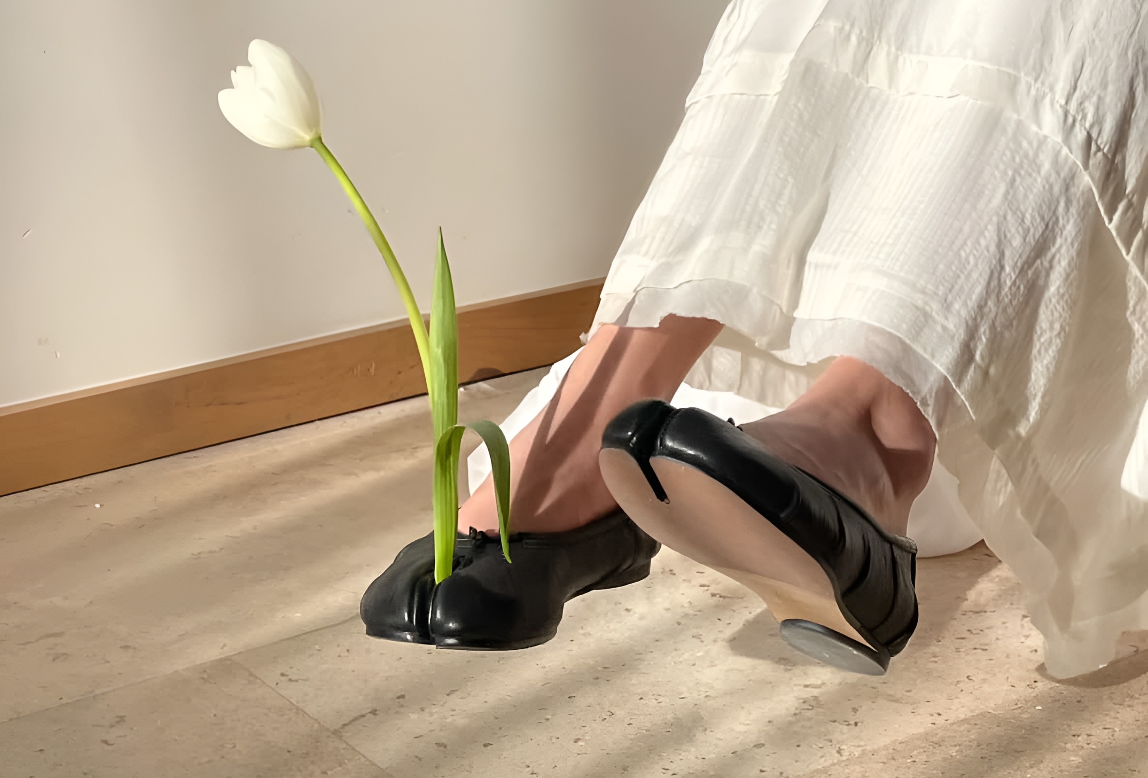 White tulip growing through black shoes on tan floor.