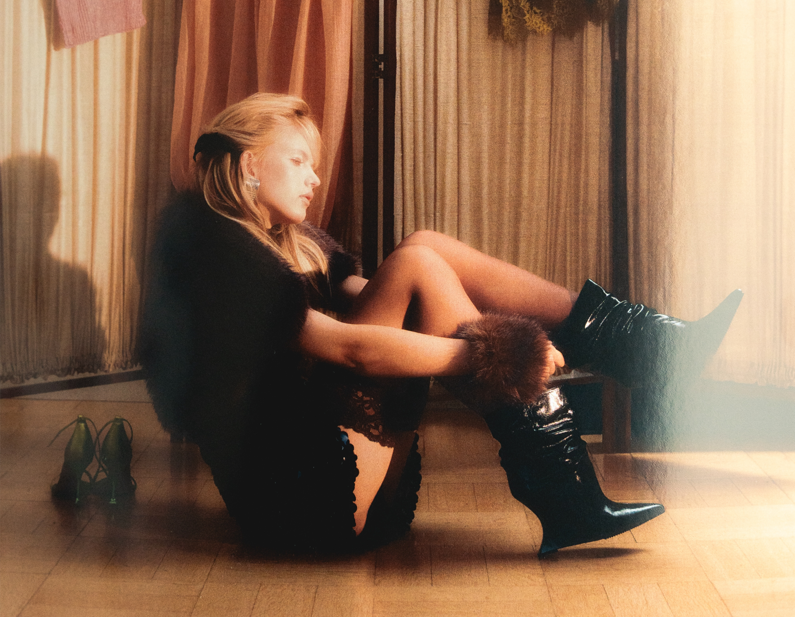 Woman in fur-trimmed outfit and boots sitting on a wooden floor, surrounded by curtains.