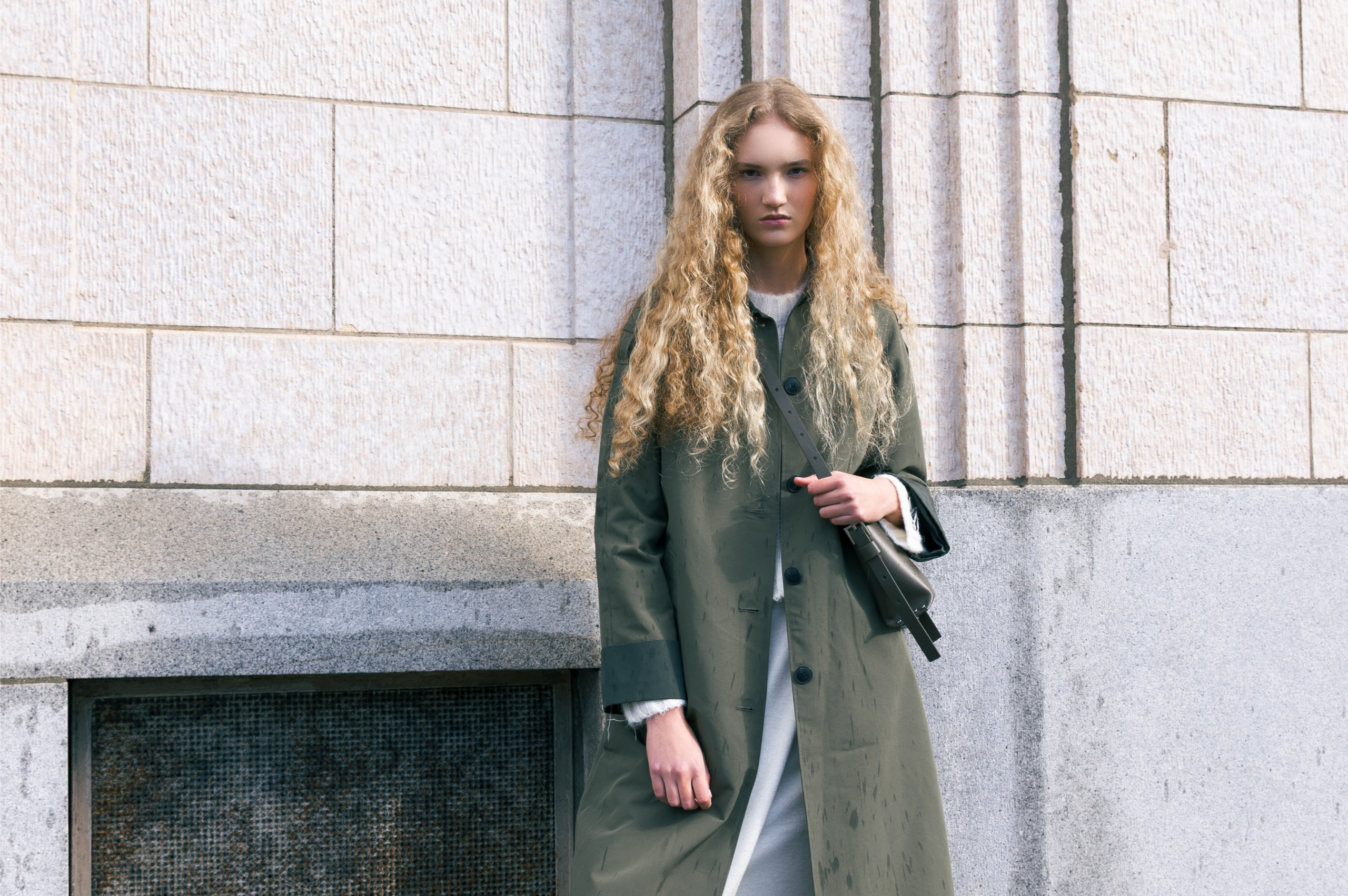 Woman with curly hair in a trench coat standing against a stone wall.