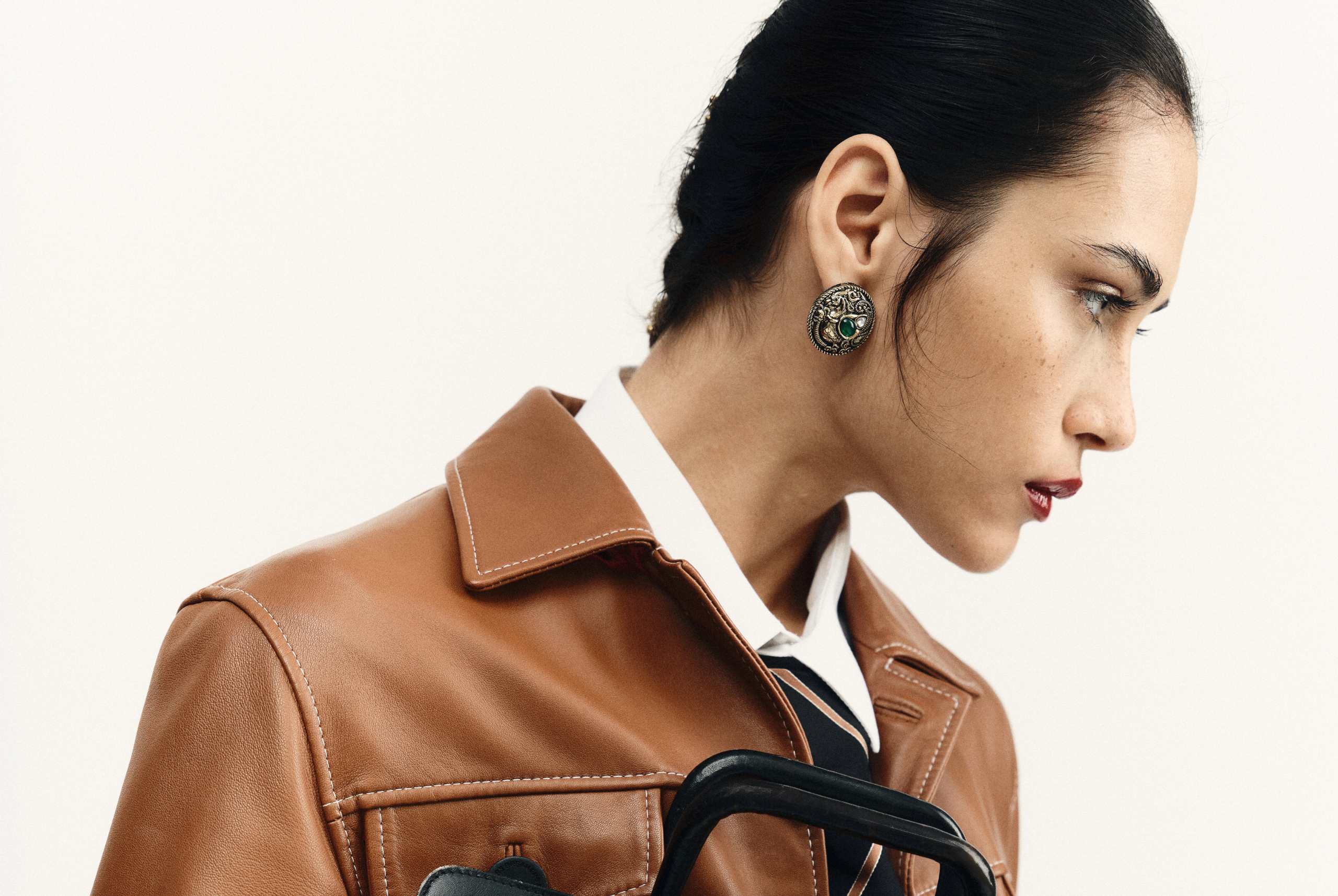 Woman in a brown leather jacket with ornate earrings, looking sideways.