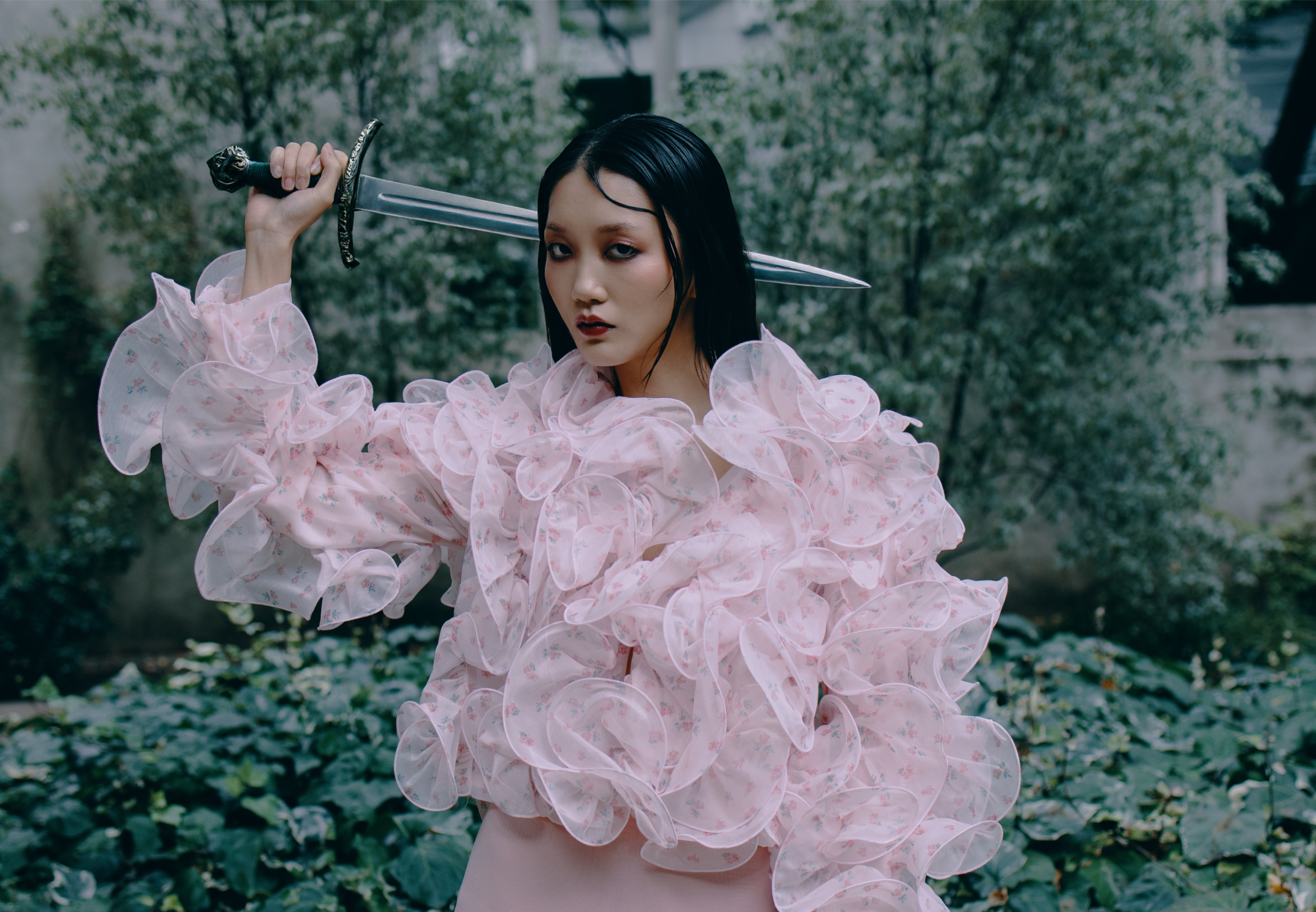 Woman in ruffled pink blouse holding sword in garden backdrop.