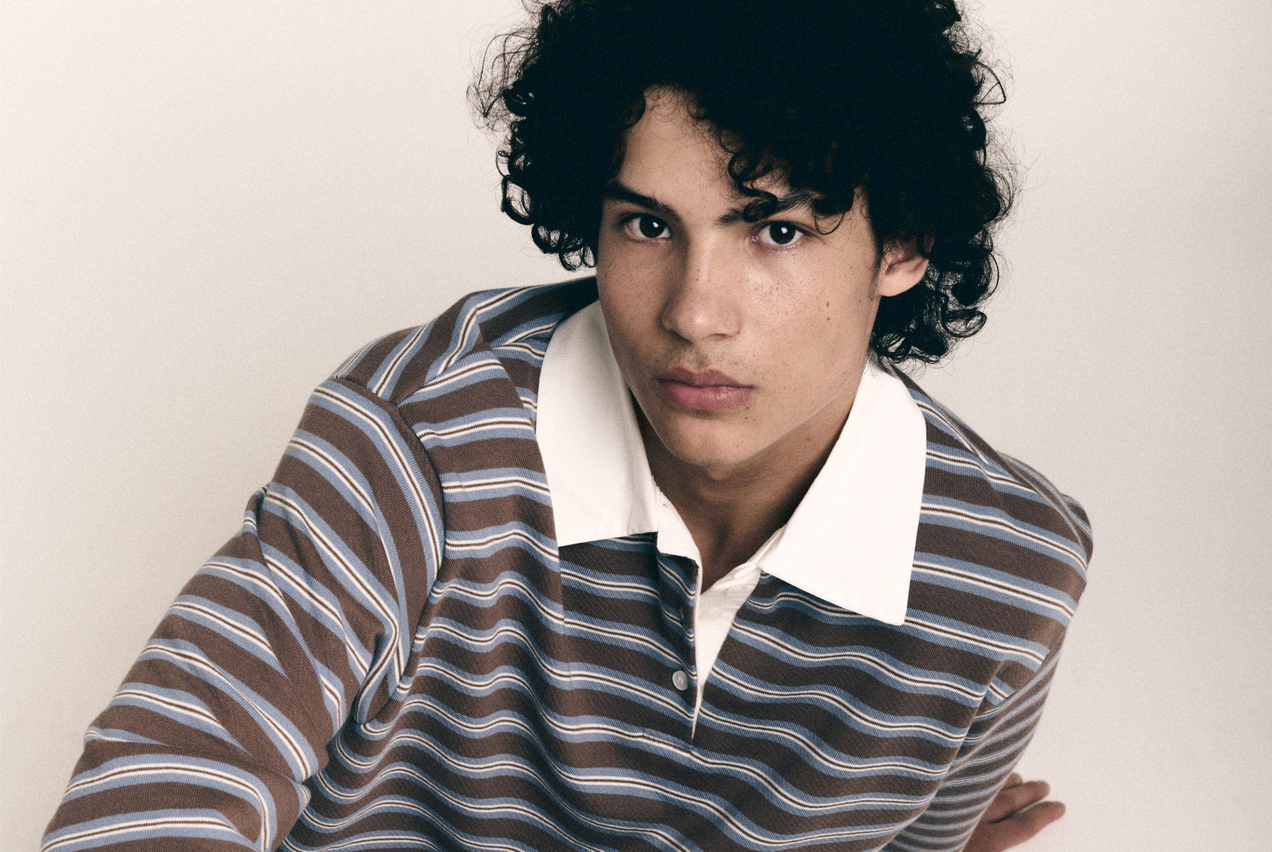 Person in brown striped shirt with white collar, curly hair, neutral expression.