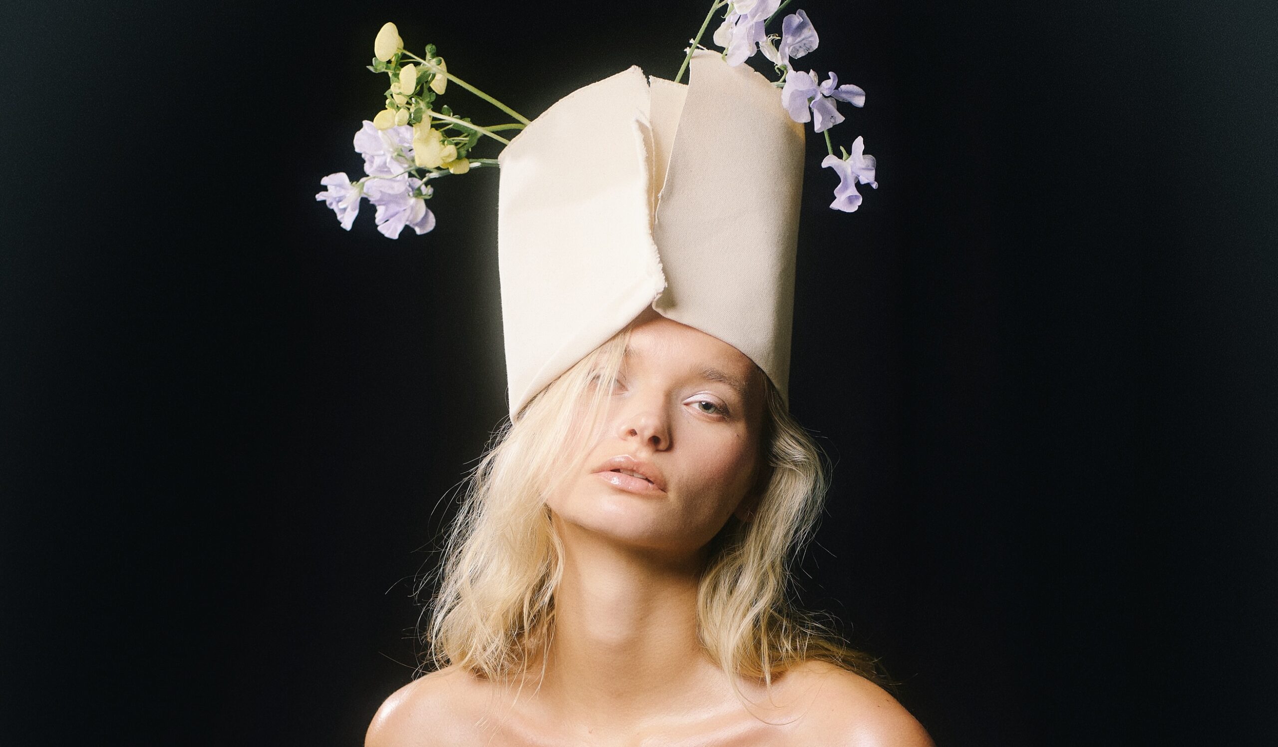 Model wearing artistic paper hat with flowers against a dark background.