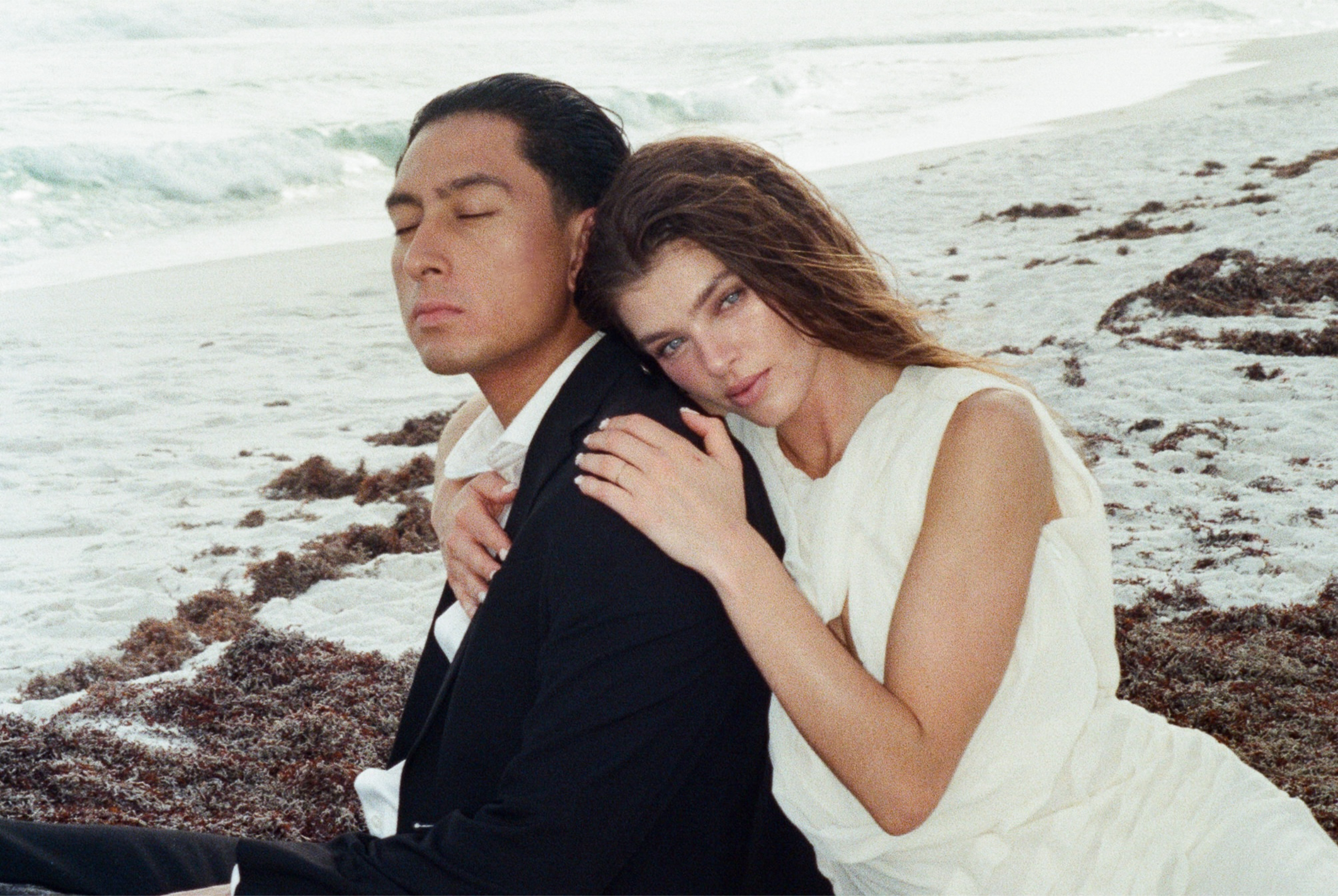 Couple embracing on a serene beach, dressed in elegant outfits.