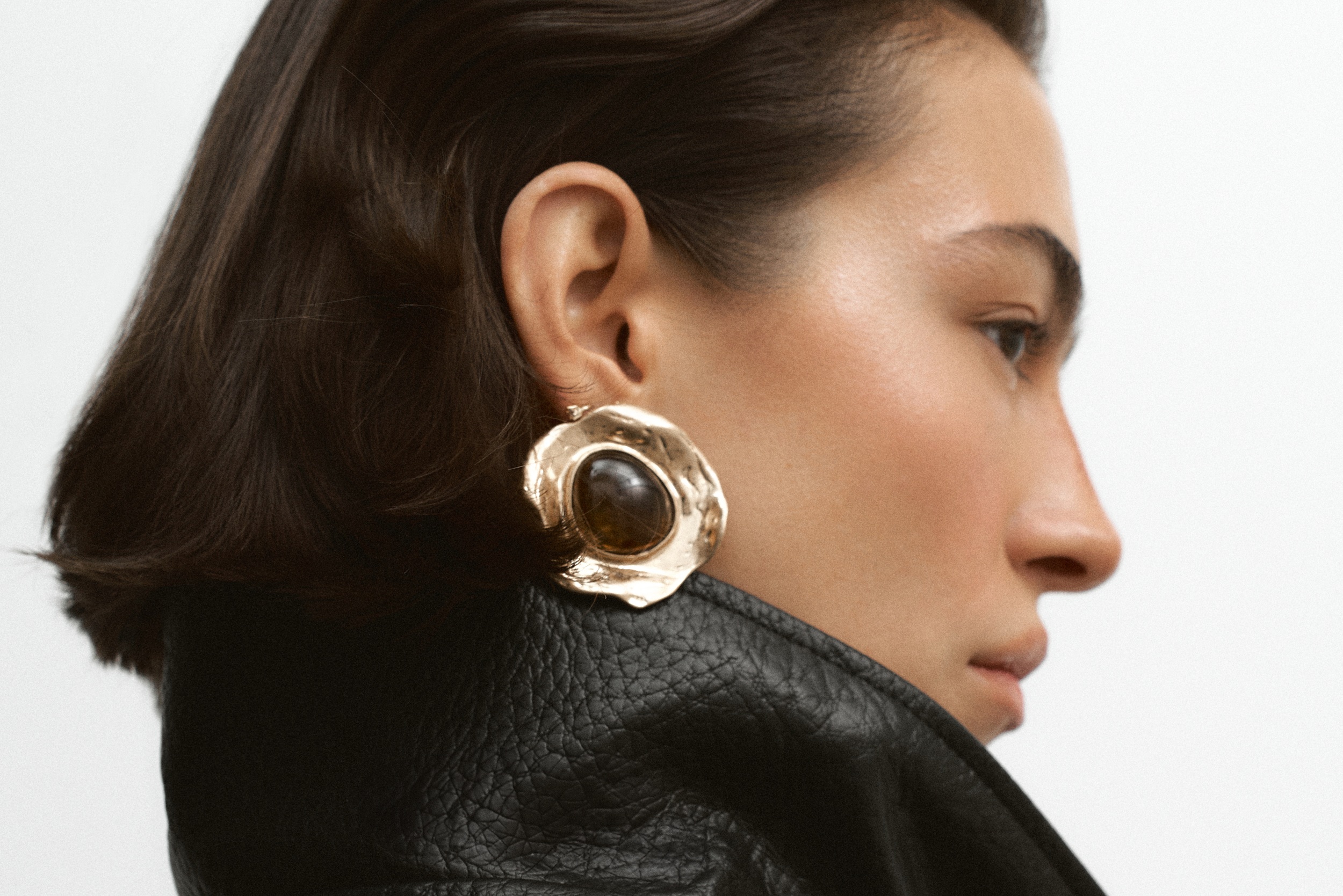 Woman wearing oversized gold earring and leather jacket, side profile view.