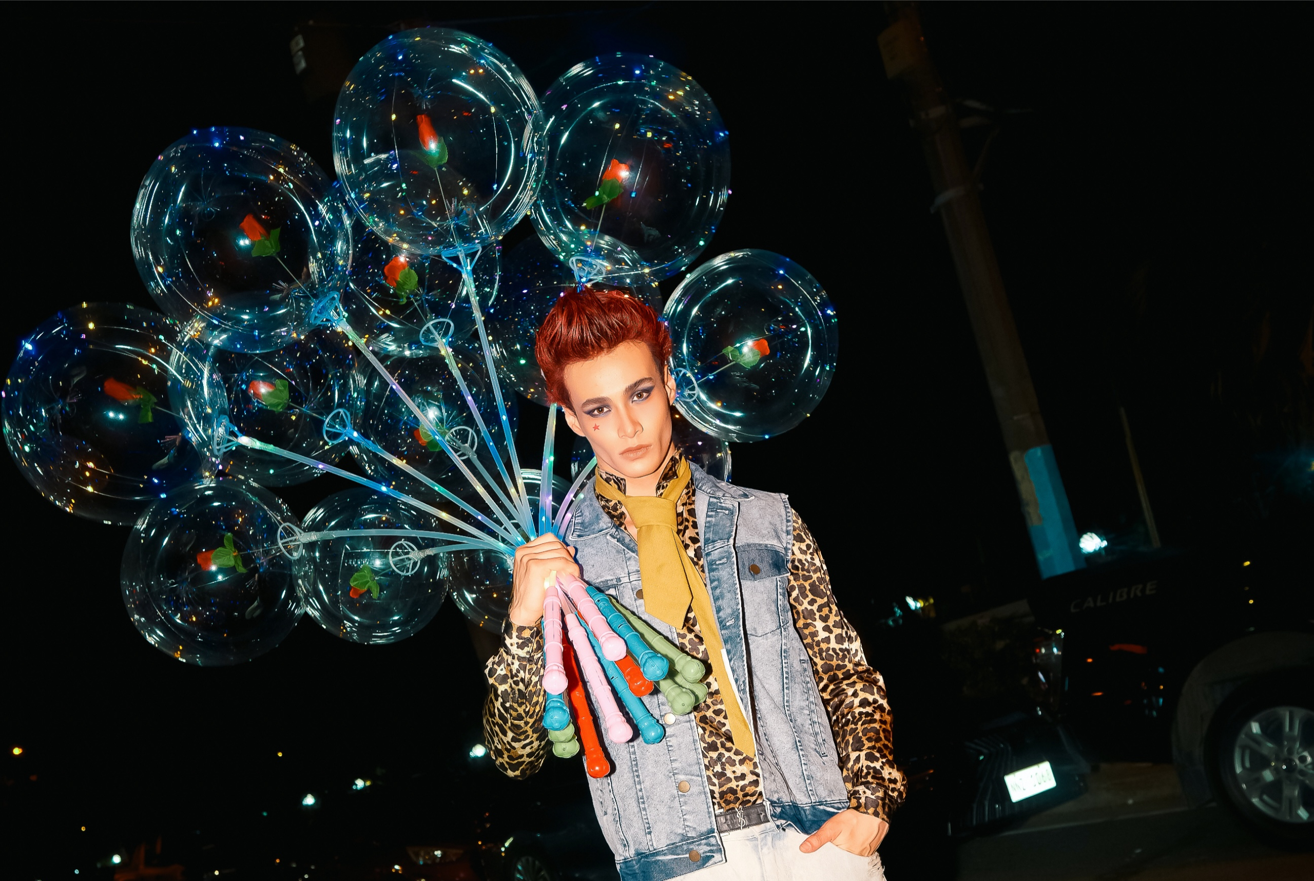Man holding glowing balloons at night, wearing stylish denim and leopard print.