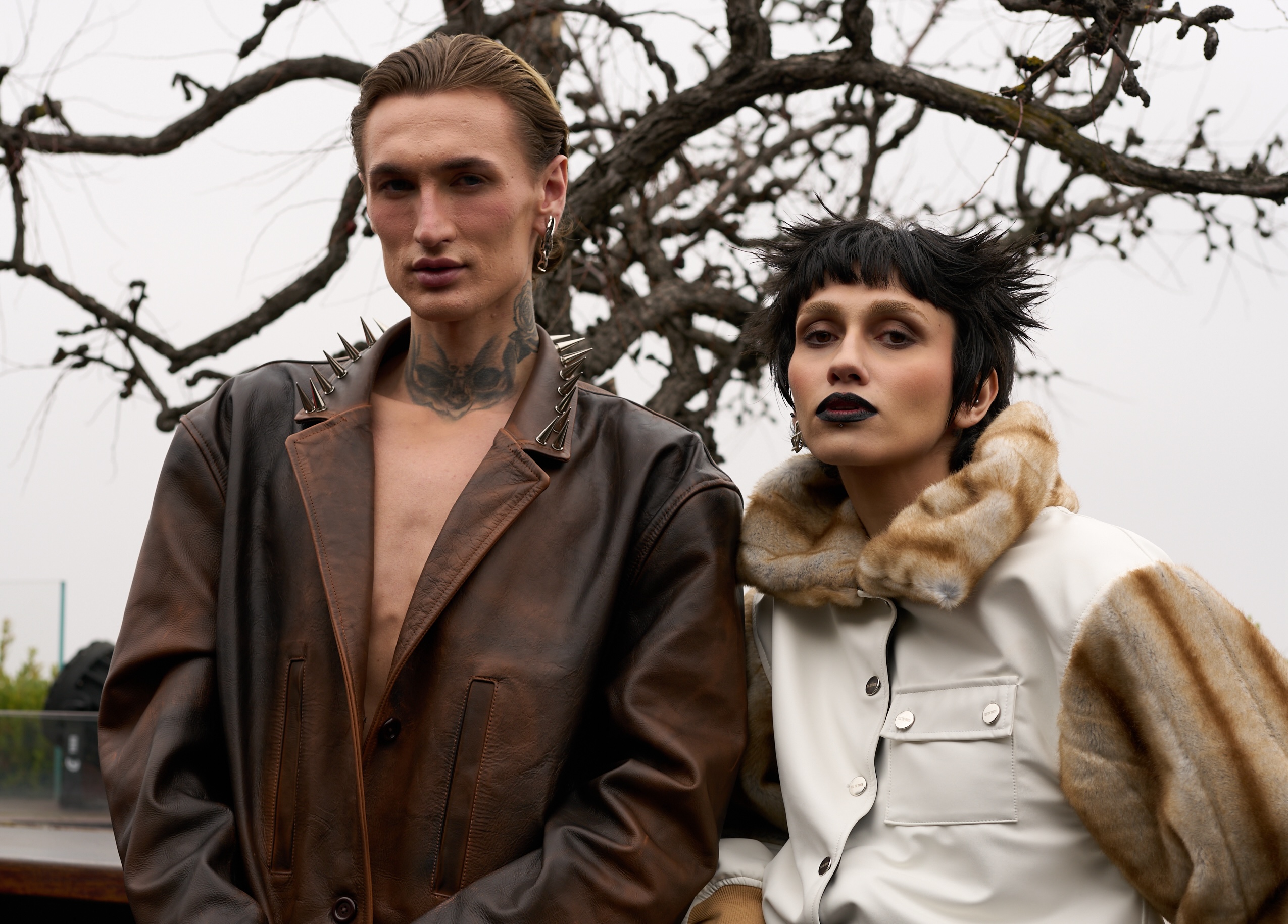Two models in edgy fashion outfits pose outdoors with bare trees in the background.