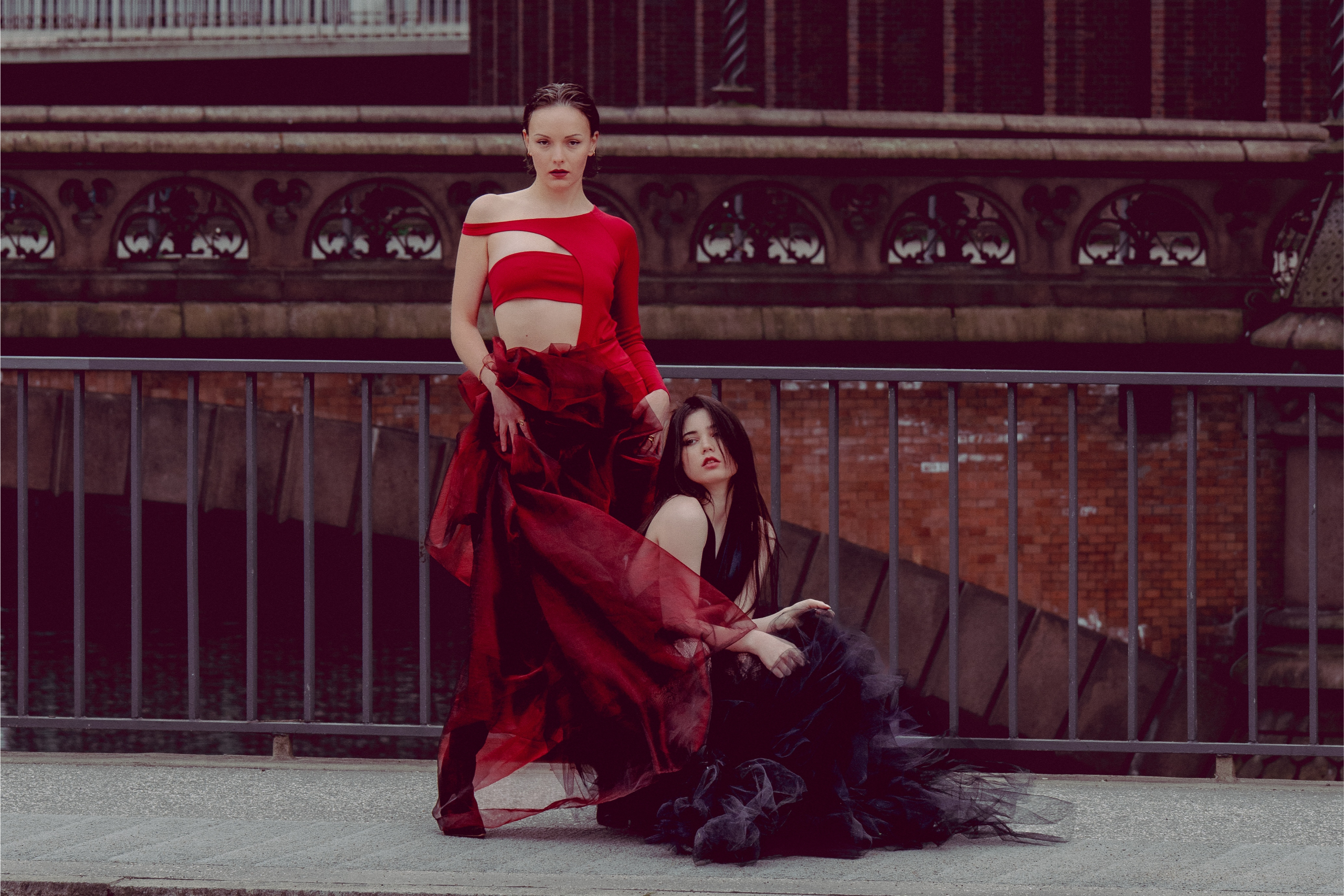 Two models in elegant red and black dresses by an ornate urban bridge.