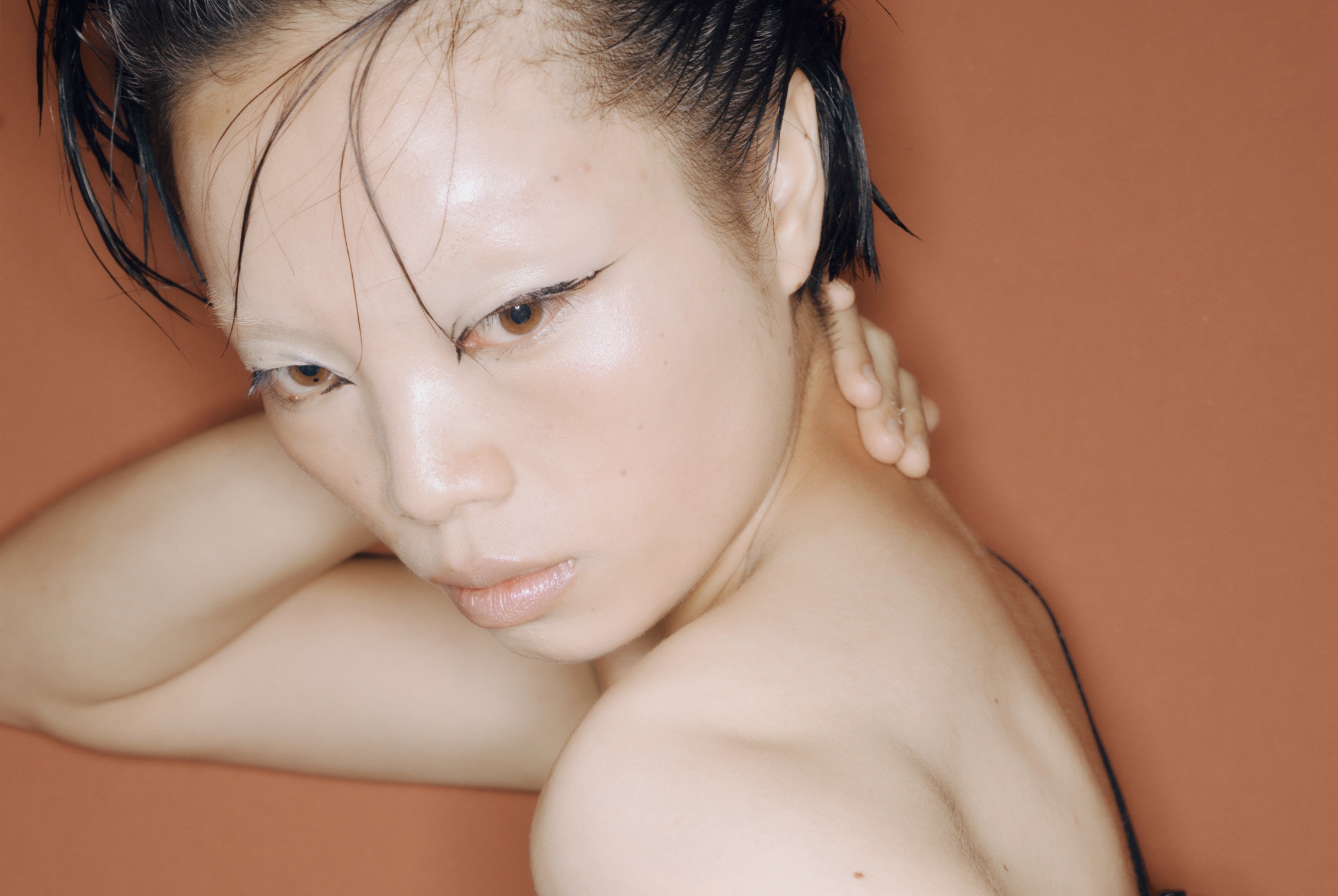 Close-up of a model with wet-look hair and dramatic eyeliner, against a coral background.