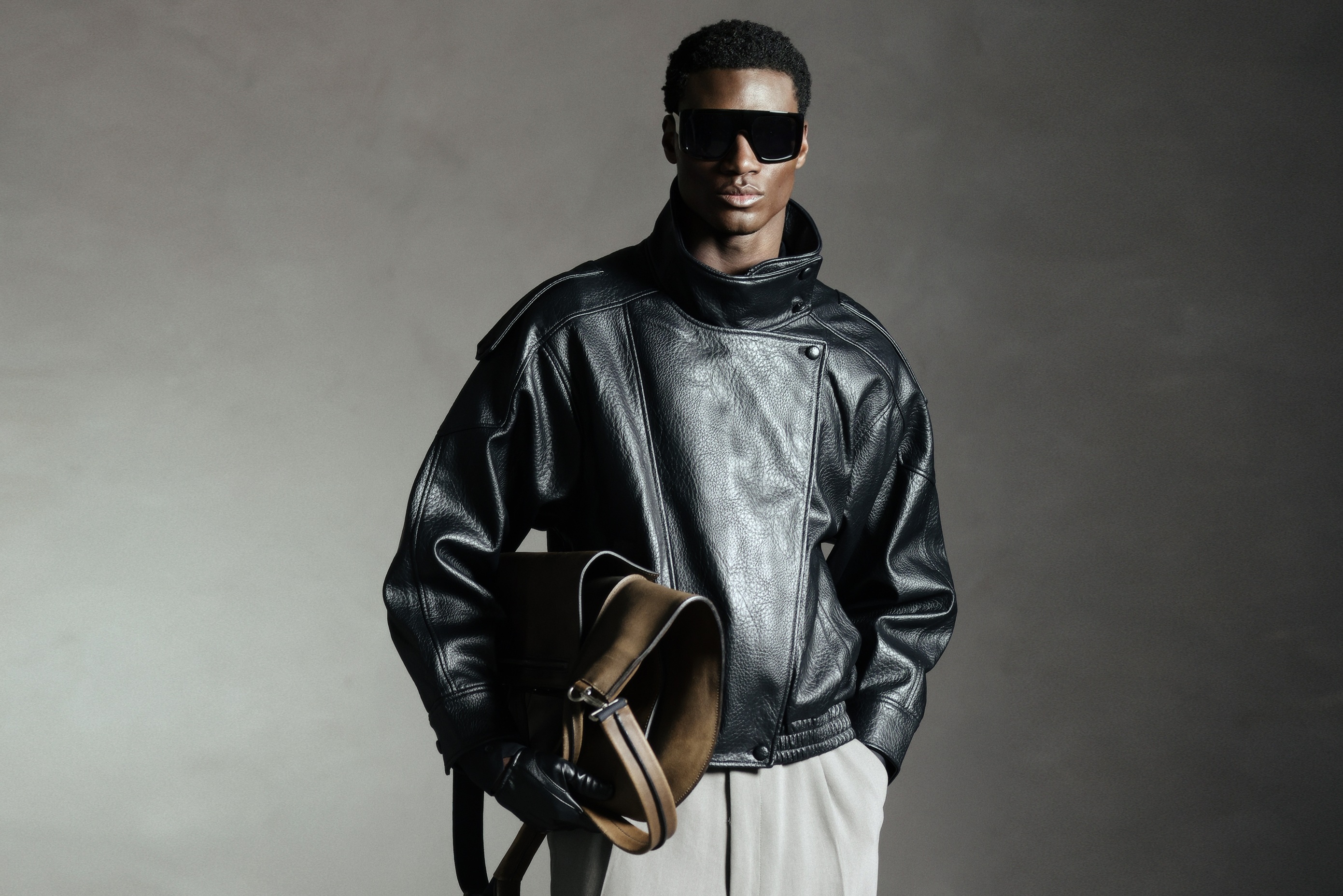 Man in black leather jacket with sunglasses holds brown bag against gray background.