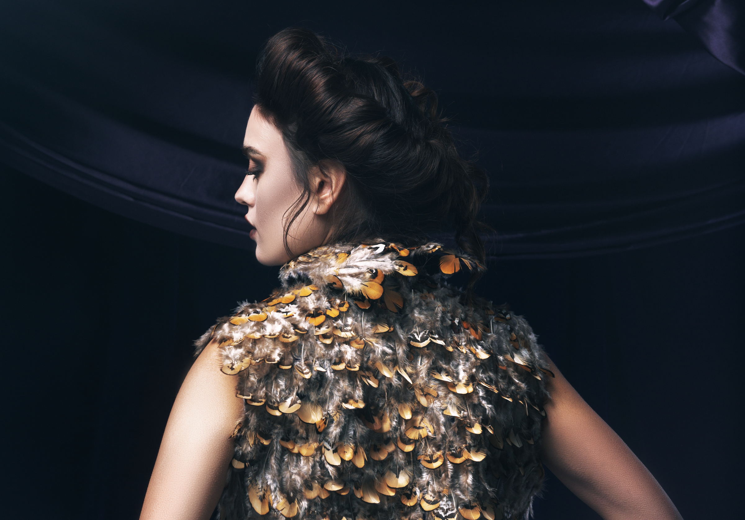 Woman with intricate updo wearing a feather-embellished garment against dark backdrop.