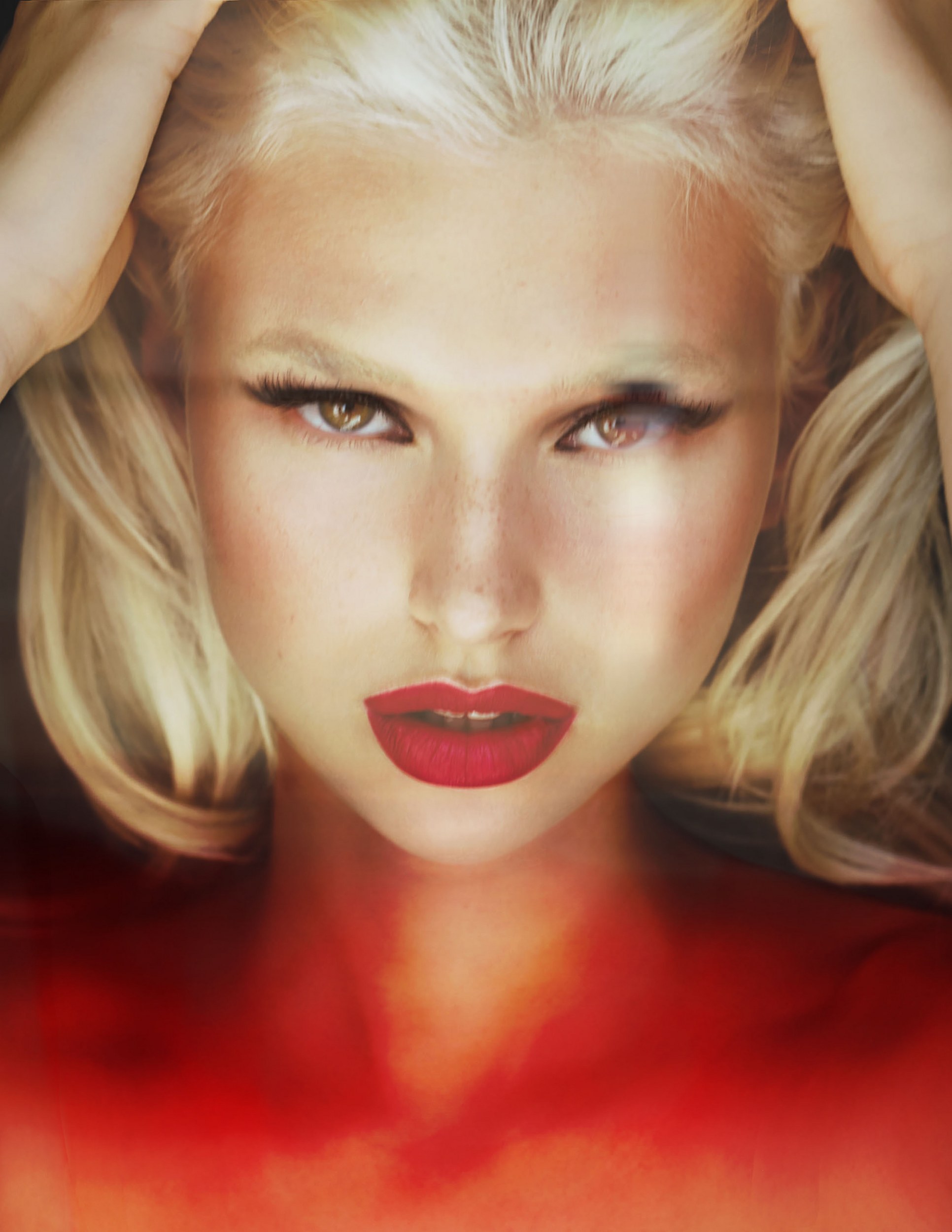 Blonde woman with red lips and intense gaze, hands in hair, artistic lighting.