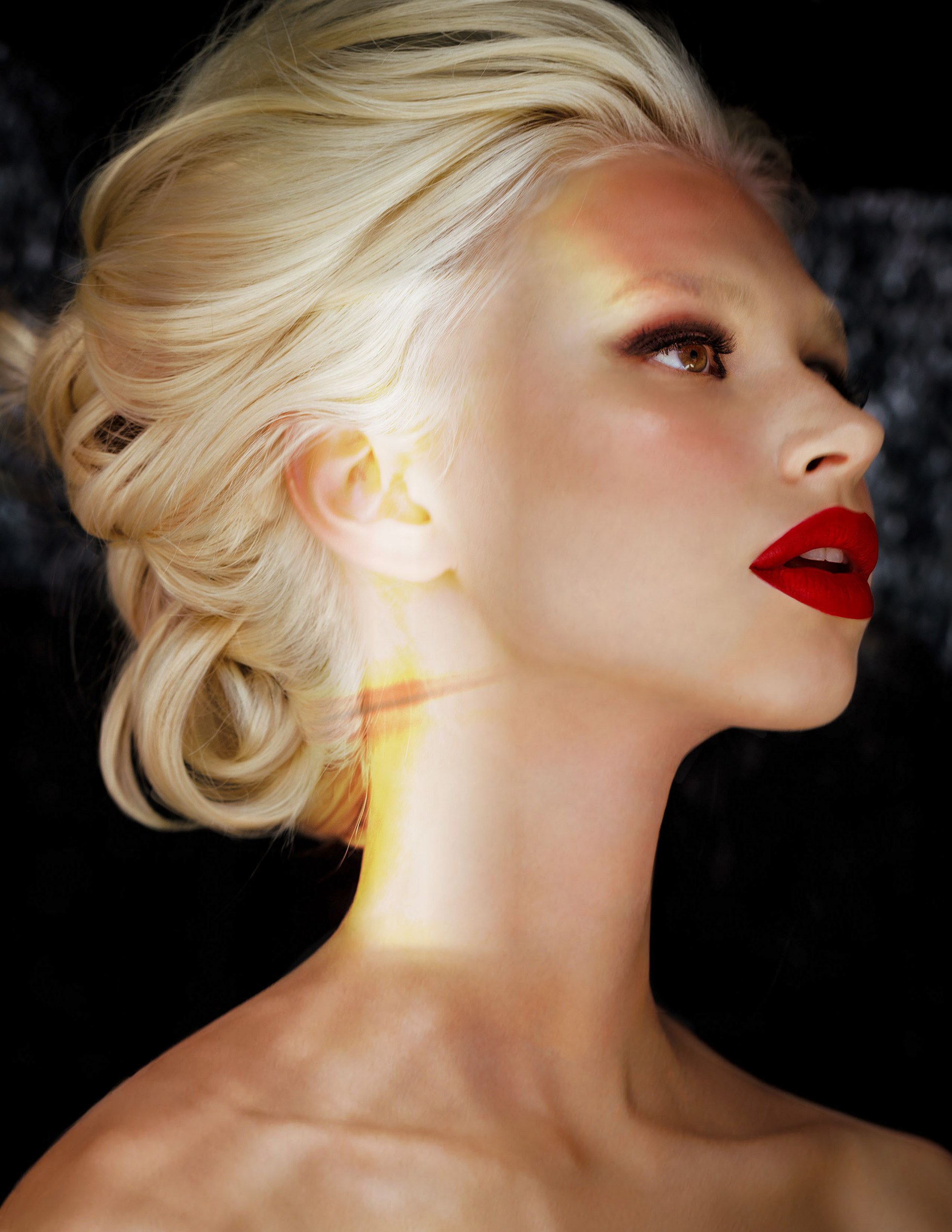 Blonde woman with red lips and elegant updo in profile against dark background.