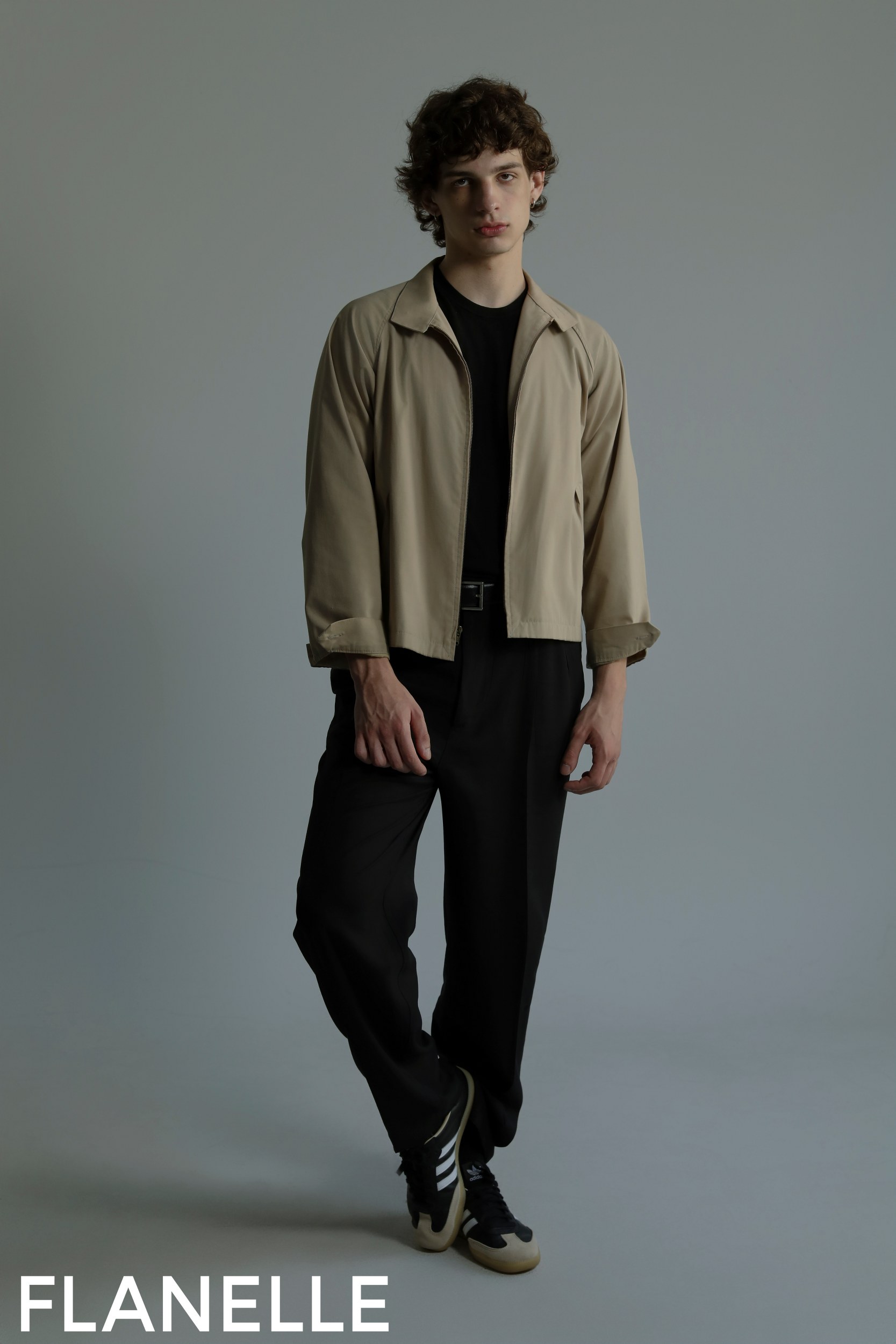 Model in beige jacket and black pants by Flanelle, standing in a minimalist pose.