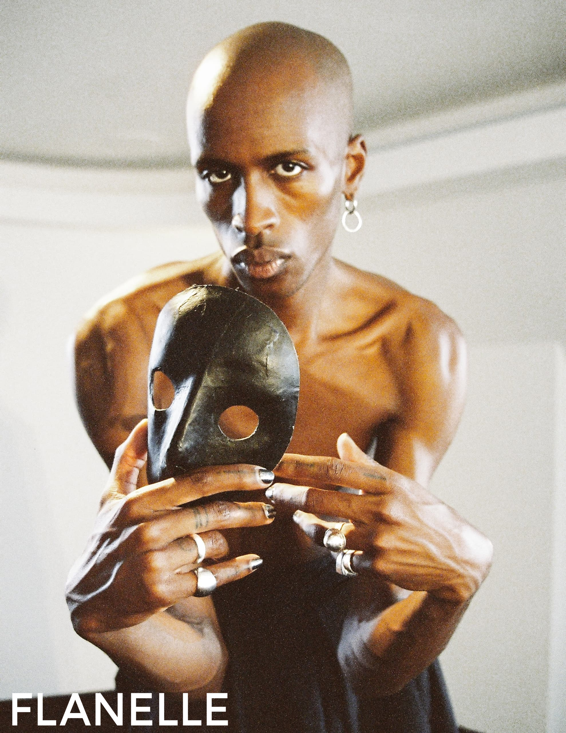 Person holding black mask, rings adorning fingers, dramatic expression in artsy setting.