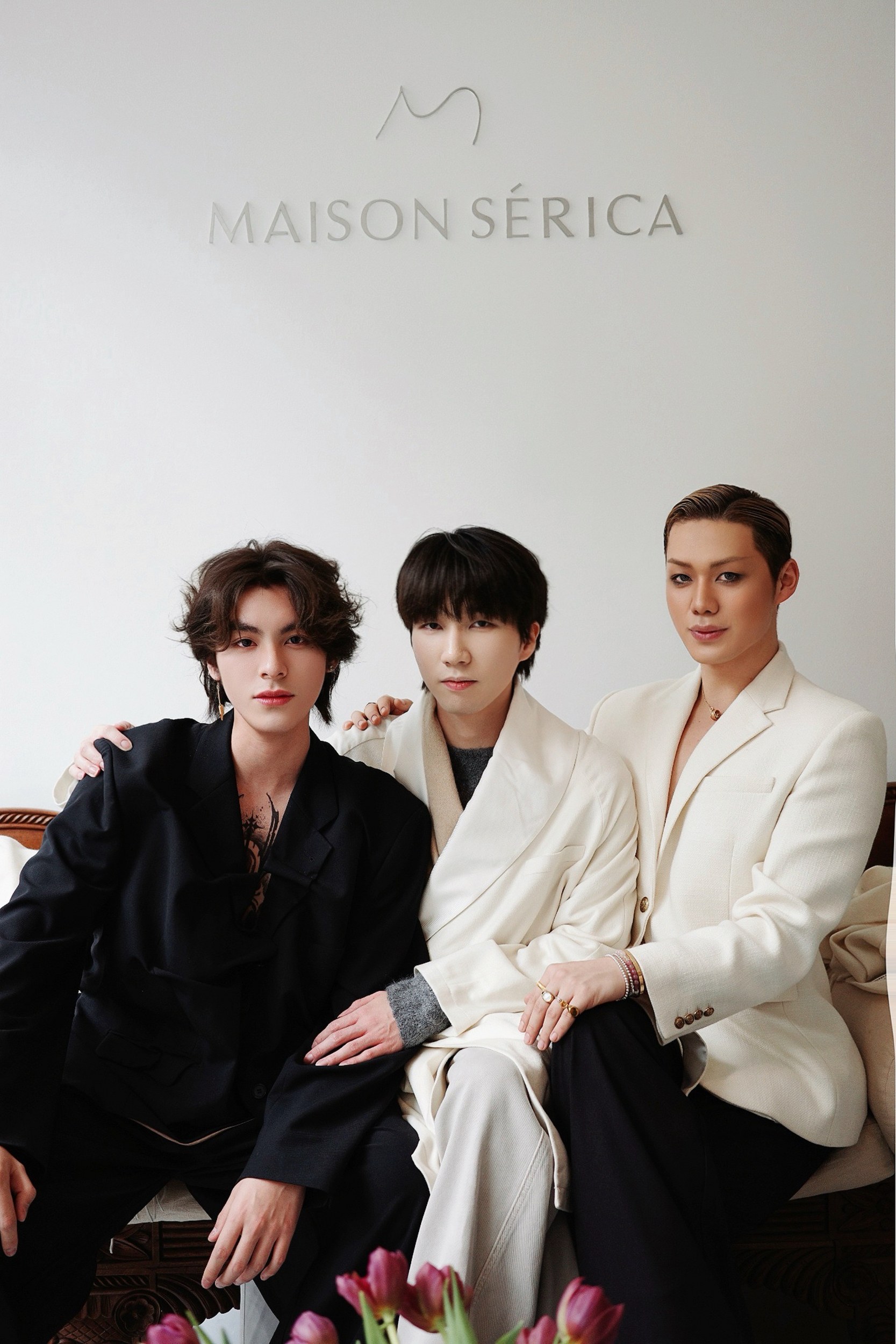 Three people in stylish suits at Maison Sérica, sitting on a sofa with tulips in view.