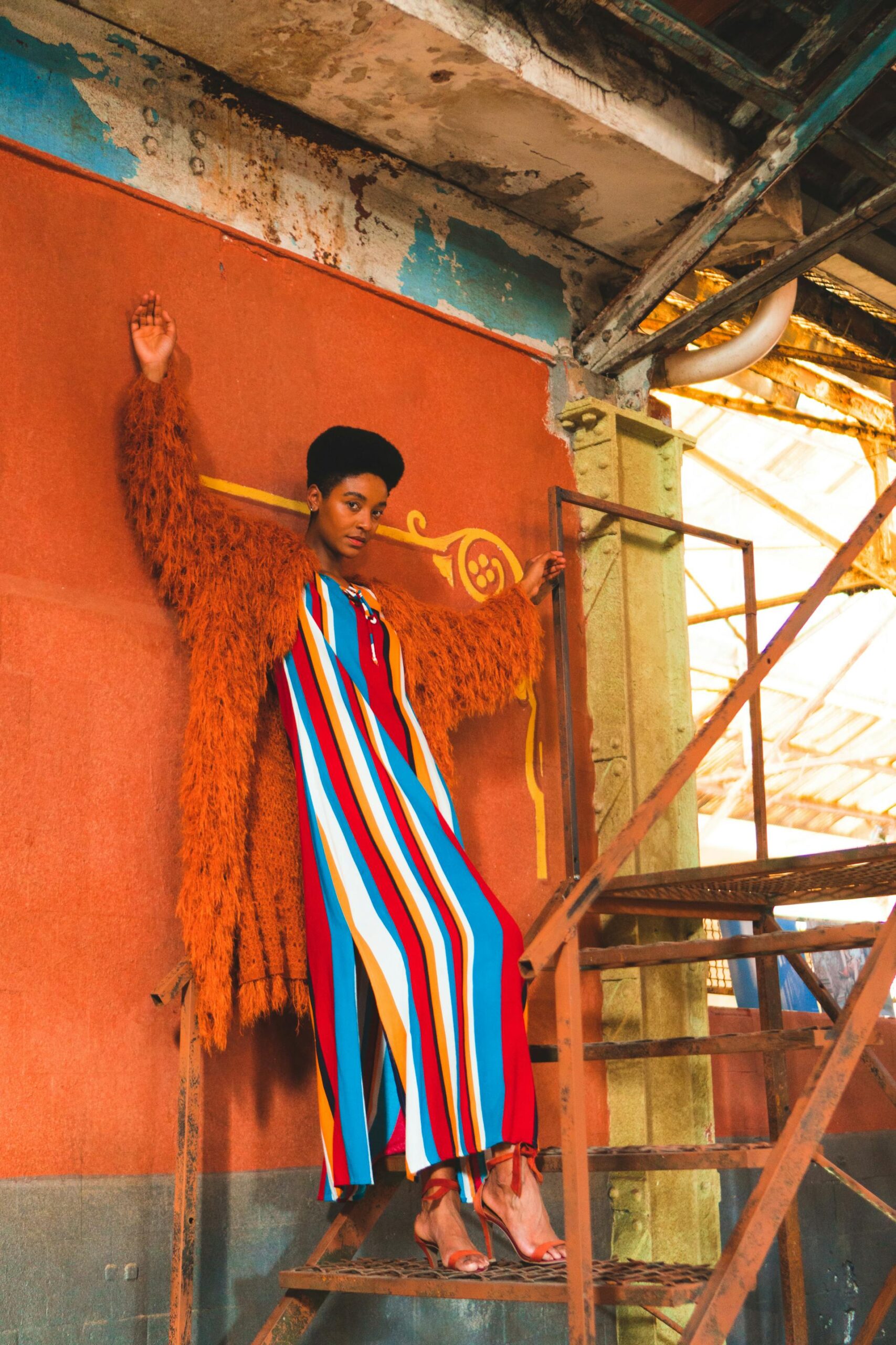 Model in vibrant striped dress and orange coat on industrial staircase.