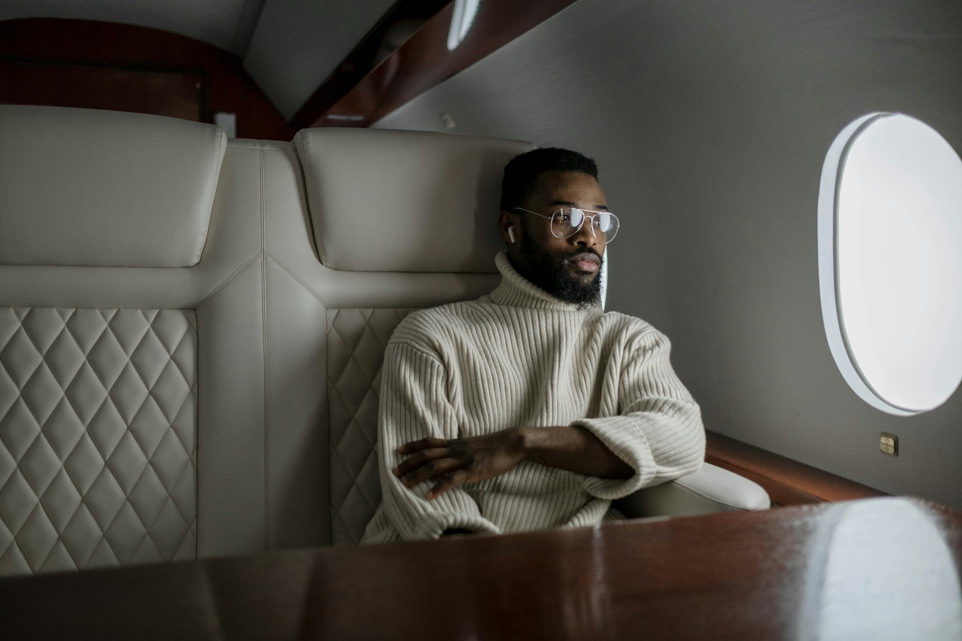 Man relaxing in private jet, wearing glasses and sweater, looking out window.