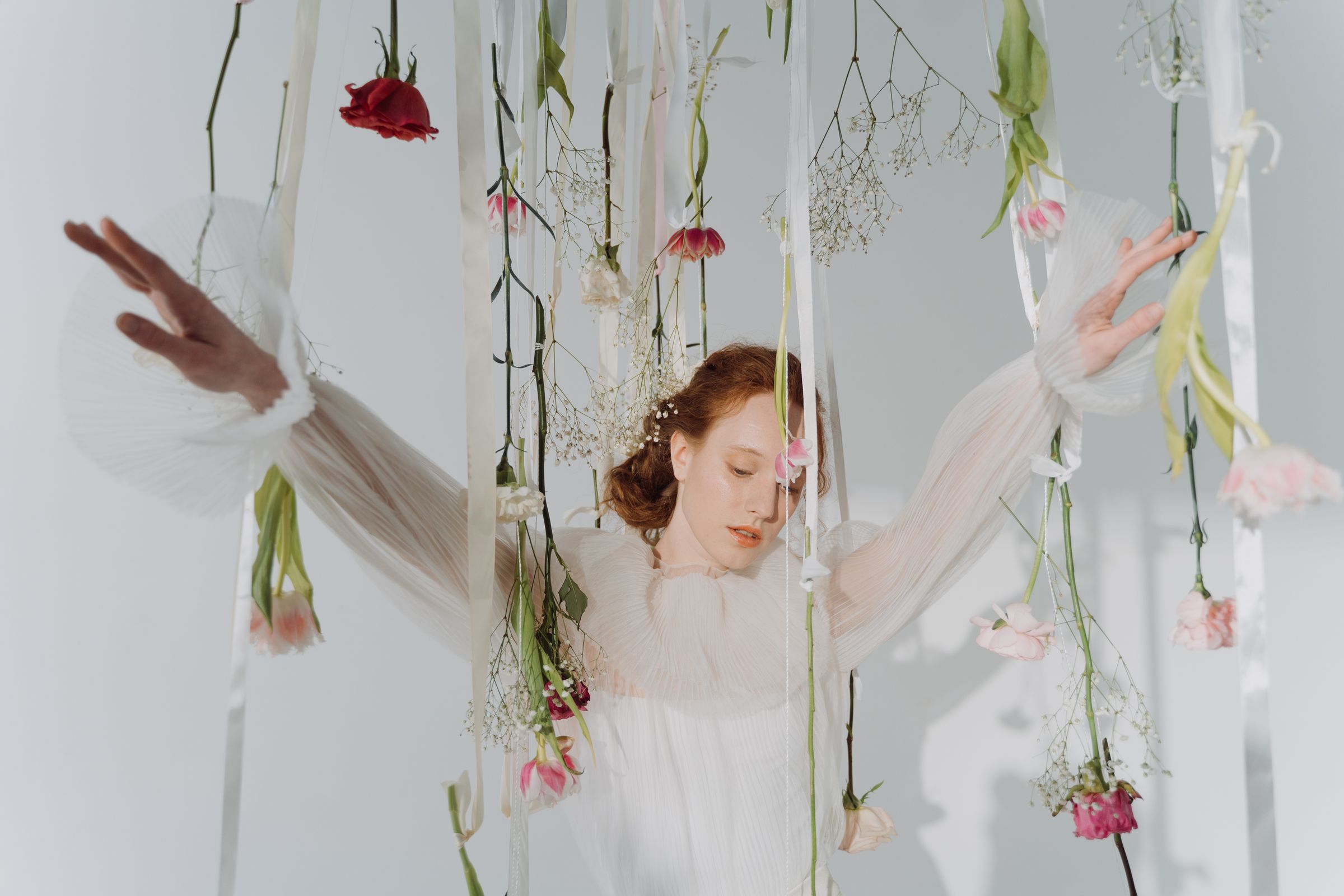 Woman in a white dress surrounded by hanging flowers and ribbons.