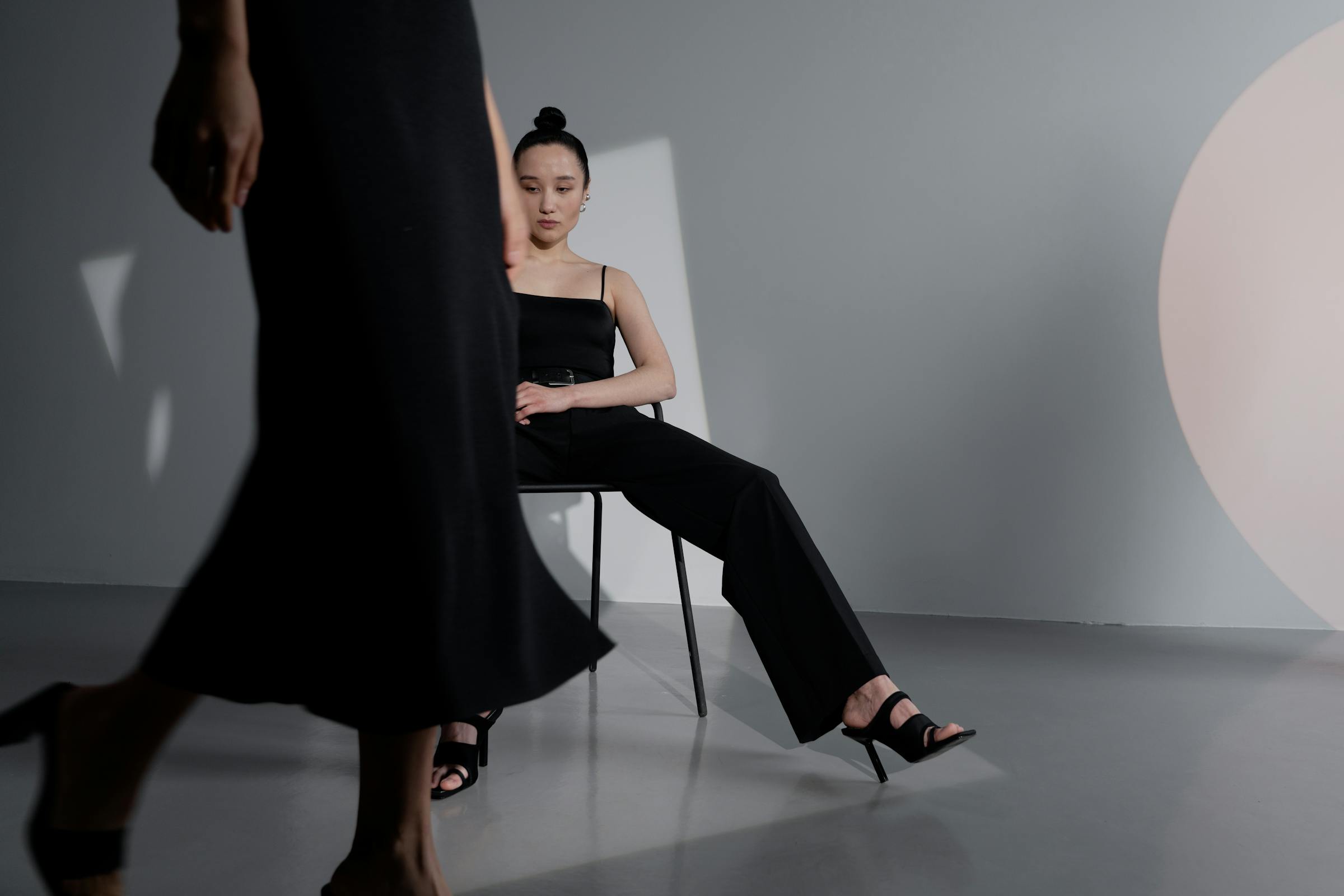 Model sitting in black outfit with another walking by, minimalist studio setting.
