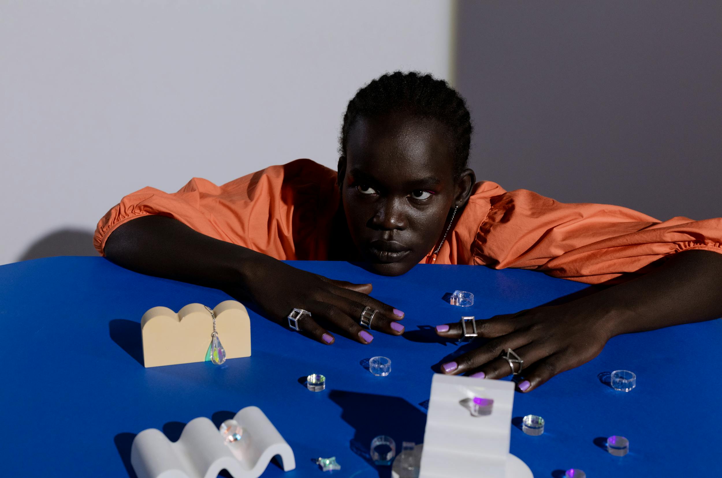 Fashionable display of jewelry; model in orange against blue table with rings and decor.