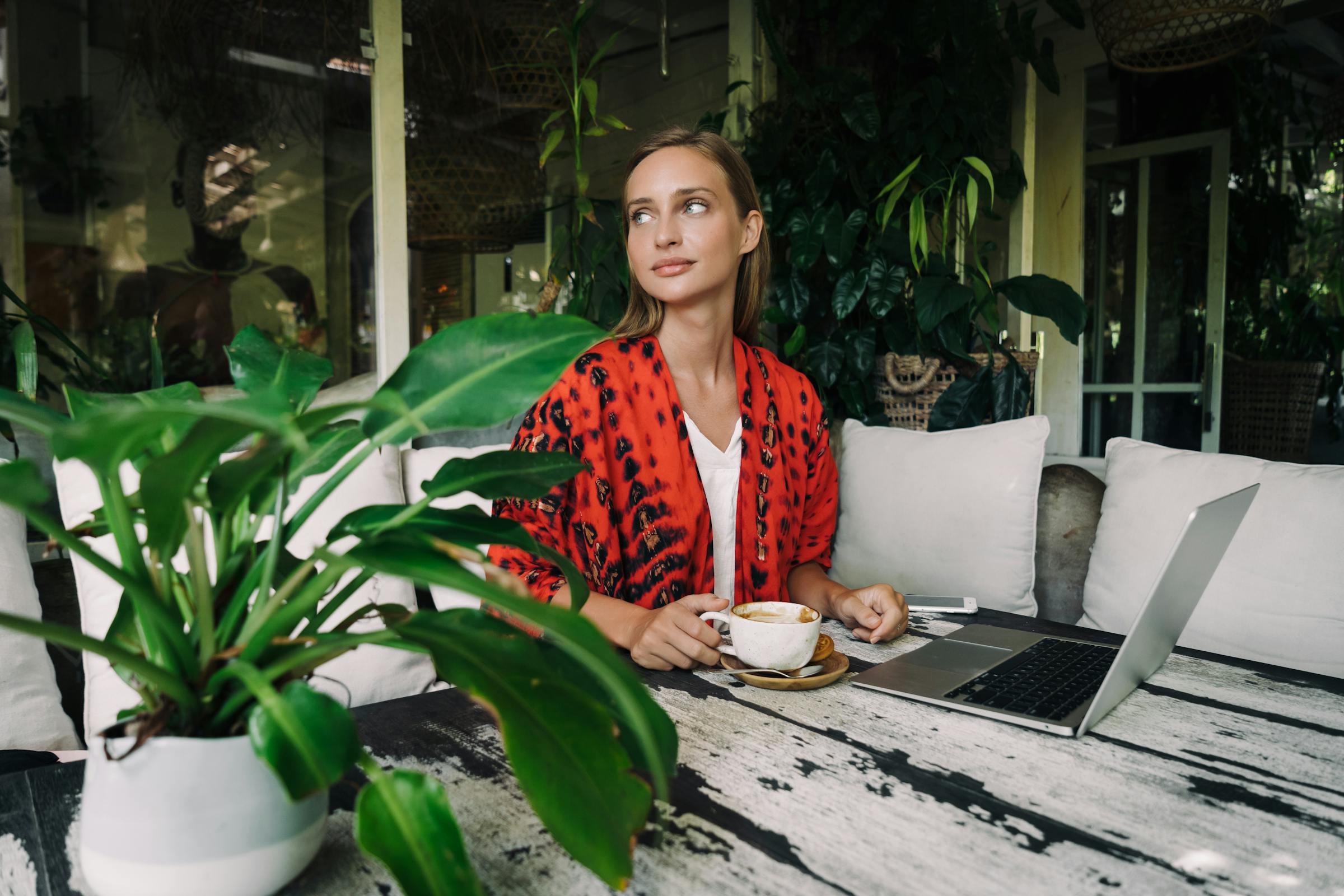 Woman enjoying coffee at a cafe patio with a laptop, surrounded by lush greenery.