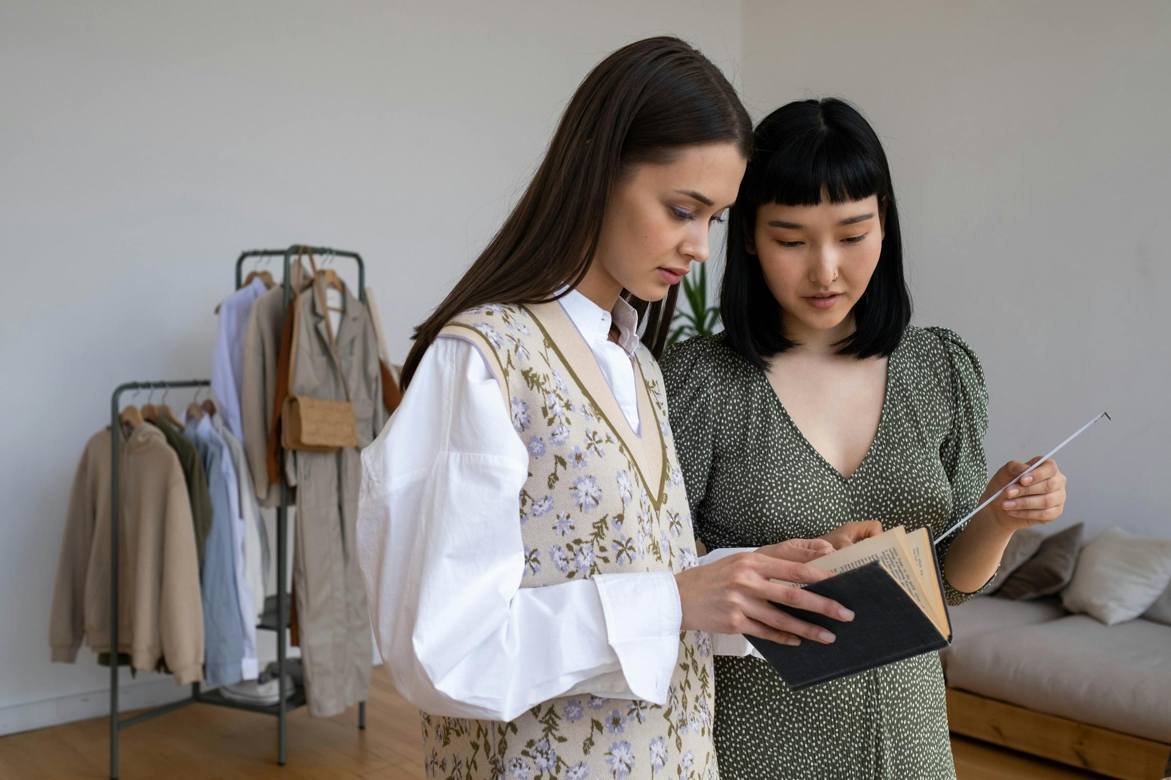 Two women reading a book in a stylish room with clothing rack in the background.