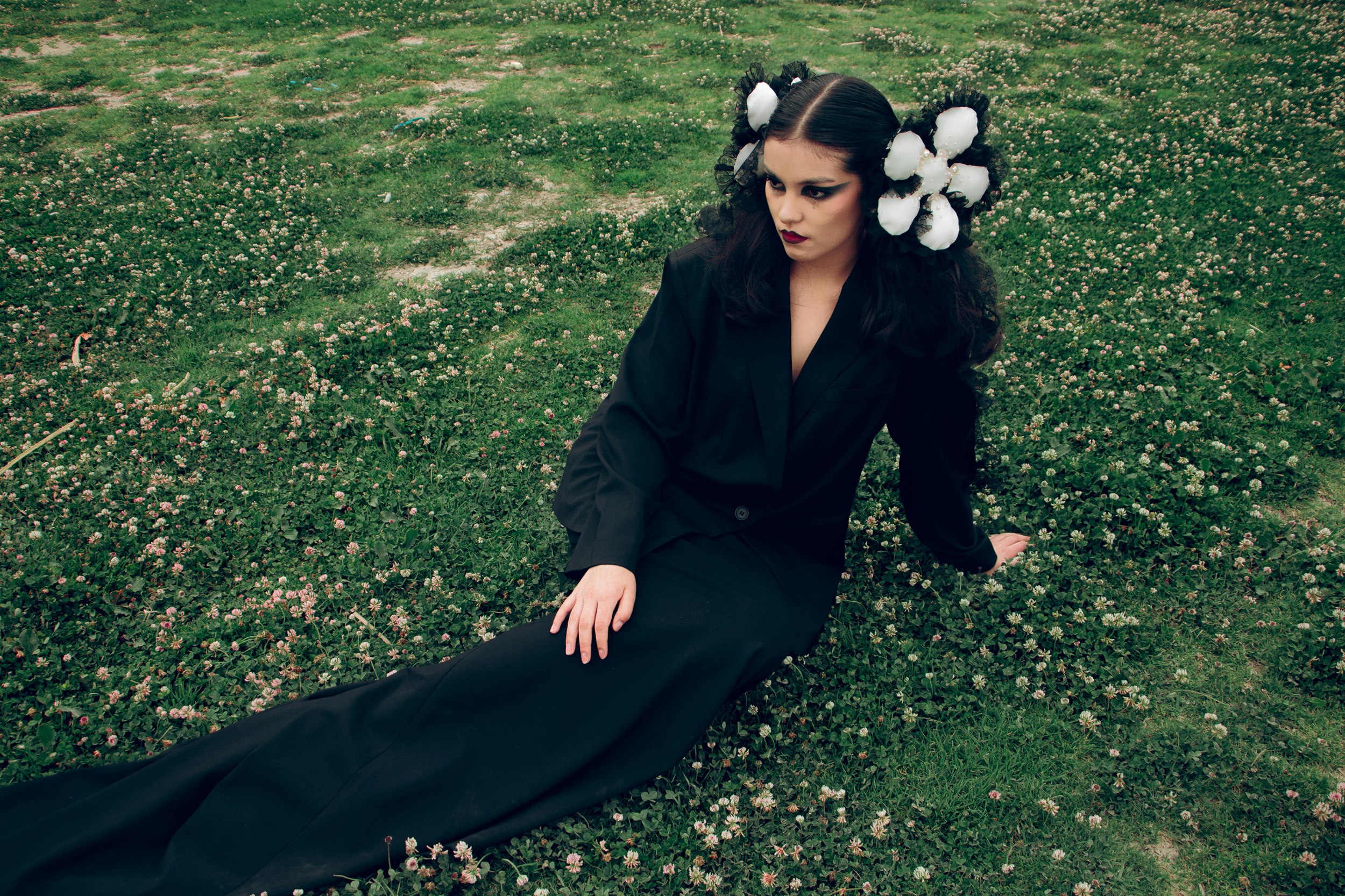 Woman in black suit with floral hairpiece sitting on grass.