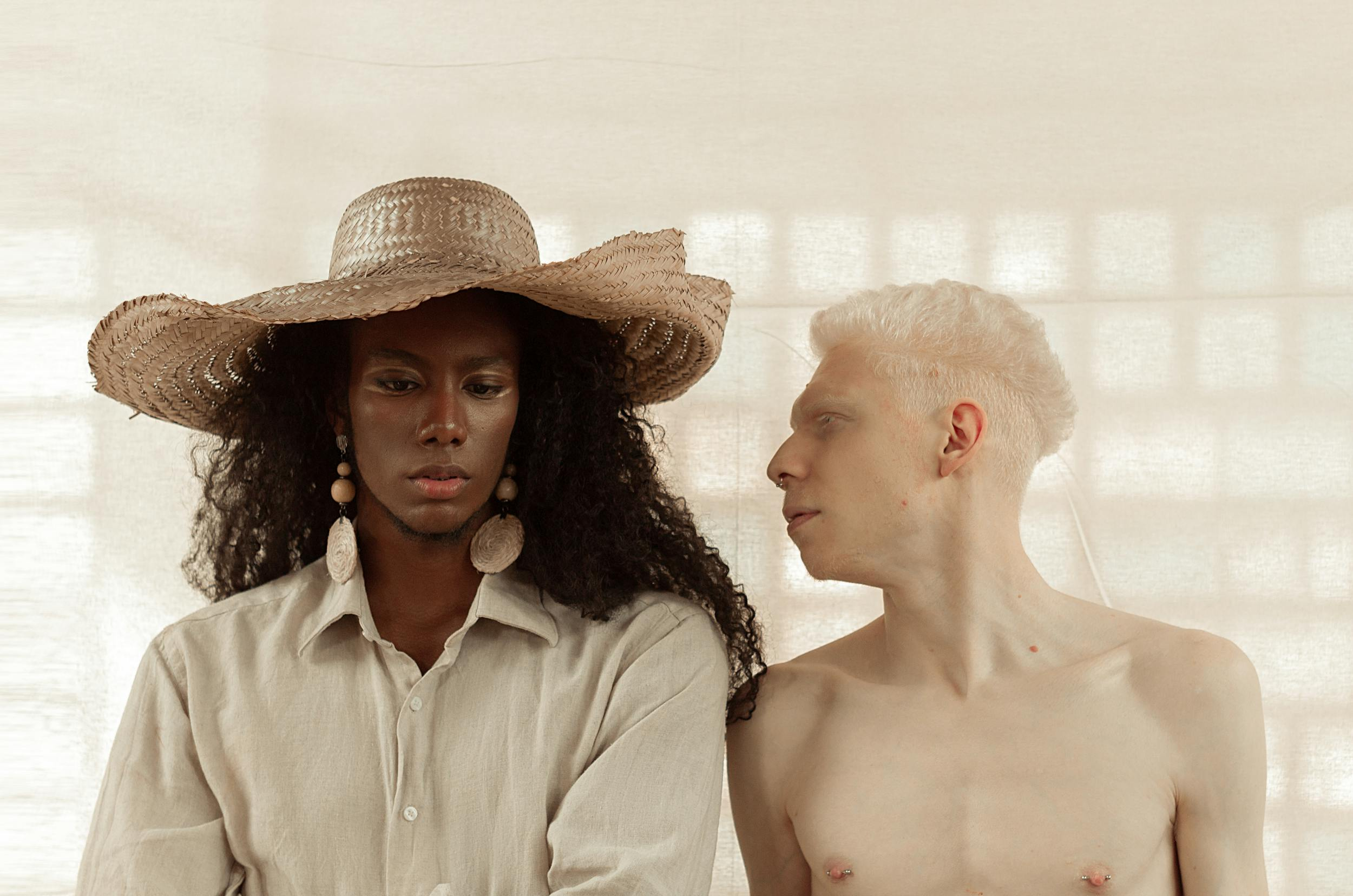 Two individuals in contrast, one in a straw hat and the other shirtless, against a soft backdrop.